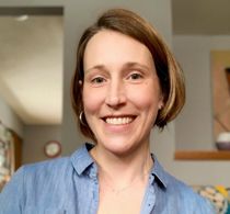 Headshot of a smiling woman with short brown hair wearing a blue shirt, standing indoors in a softly lit room. The atmosphere is warm and friendly.